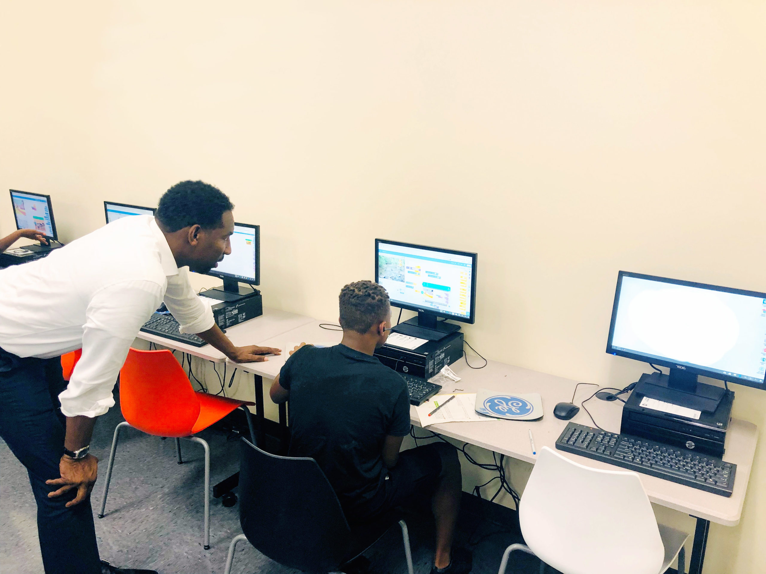 A young Black man & @Promise student is seated at a workstation. An instructor leans on the desk and looks over the student's shoulder. The instructor is a Black man of average build in his mid-thirties. He is wearing a white dress shirt with the sleeves rolled up.