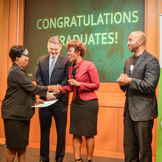 Presenters handing a diploma to a TechBridge Technology Career Program graduate. A middle-aged Black woman wearing professional dress holds her diploma in her left hand while she shakes the presenter's hand with her other.