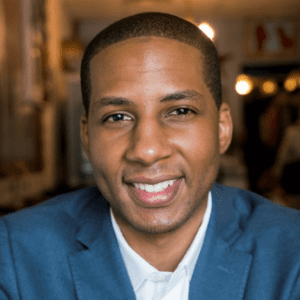 Informal portrait of John DeShazer. He is a youngish Black man with short cropped hair and a big smile. He wears a blue suit jacket or sport coat and an open-collar white shirt. The blurry background suggests a restaurant or bar interior.