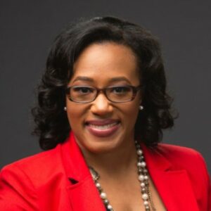 Maranie Brown studio portrait. Maranie appears a middle-aged Black woman. She has shoulder-length, dark hair and is wearing eyeglasses and a red business suit. She is also wearing a necklace featuring large wooden beads. She looks directly at the camera and has a broad, open smile.