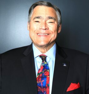 Studio portrait of Ricky Steele. Steele wears a dark business suit and an abstract patterned tie featuring saturated primary colors. He is is smiling. He has short, dark hair going gray, a broad nose, and dark eyes.