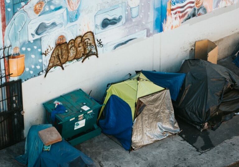 Unhoused tents on the sidewalk of Toronto street in front of a graffiti-scrawled wall.