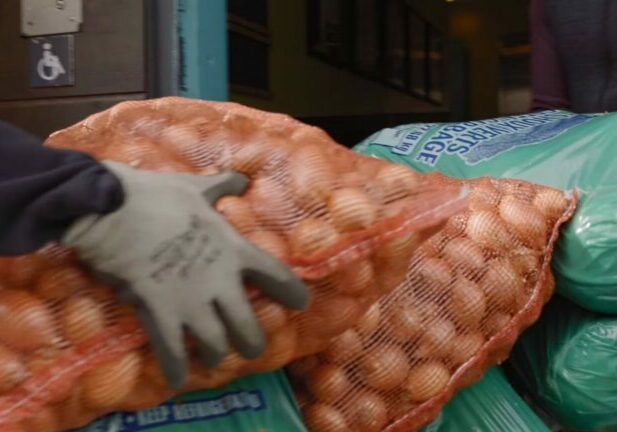 Large bags of potatoes and other produce are stacked by gloved hands.