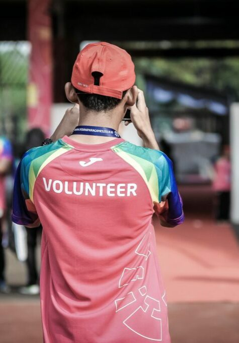 Young man taking a photograph at an event. His back is turned to the viewer. The word `Volunteeer` is emblazoned across his shirt.