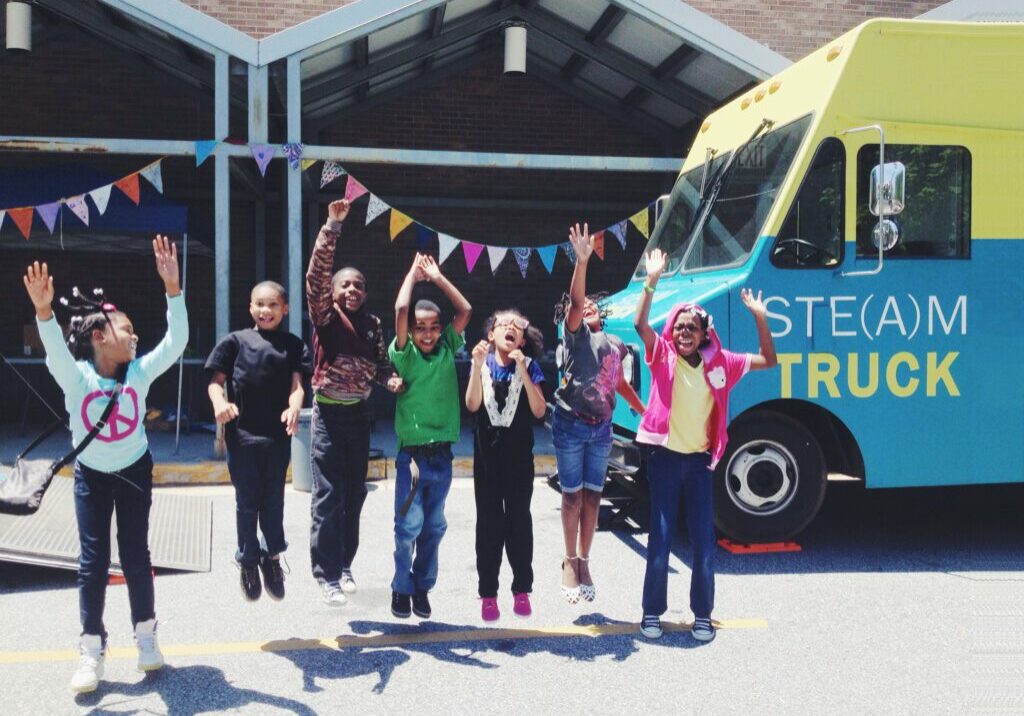 7 young Black schoolchildren jumping, waving, and smiling in front of the Atlanta Community Guilds STEM/STEAM truck